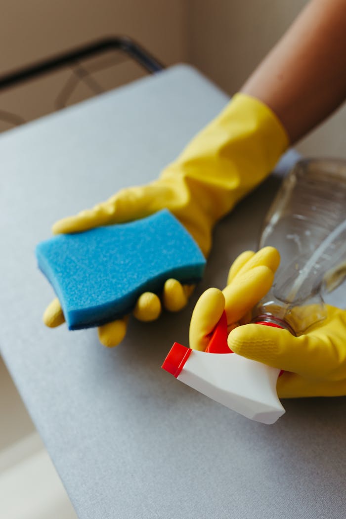 Close-up of a person cleaning a surface with a blue sponge and spray bottle, using yellow gloves.